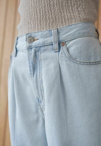 Ljusblå denimbyxor med plisserad front, försedda med en metallknapp och kopparnitar. Mjuk textur och avslappnad design.