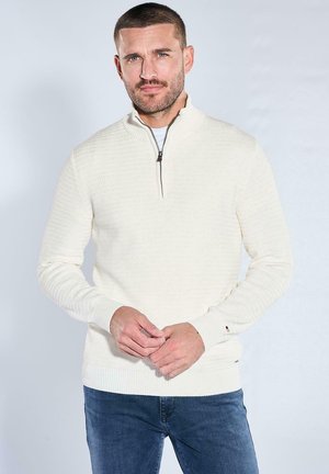 Mann mit kurzem Haar und Bart, der einen weißen, strukturierten Half-Zip-Pullover und blaue Jeans trägt und mit verschränkten Händen steht.