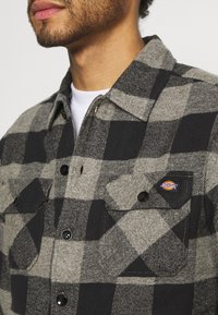 Gray and black plaid shirt made of textured fabric, featuring two chest pockets with button closures and a prominent brand logo.