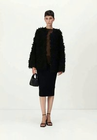 Μαύρο φουντωτό μπουφάν με μακριά μανίκια και κούμπωμα με κουμπιά, φορεμένο πάνω από καφέ τοπ και μαύρη μίνι φούστα; το μοντέλο κρατά ένα μικρό μαύρο τσάντα.