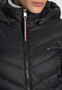 Primo piano di una persona che indossa un giaccone piumino nero con cappuccio e una zip centrale con un tirante rosso, bianco e blu.