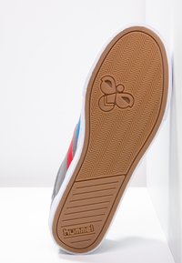 Basket grise avec des accents rouges et bleus. Dotée d'une semelle en caoutchouc avec un motif en chevron et le logo "hummel" ; semelle intermédiaire blanche.