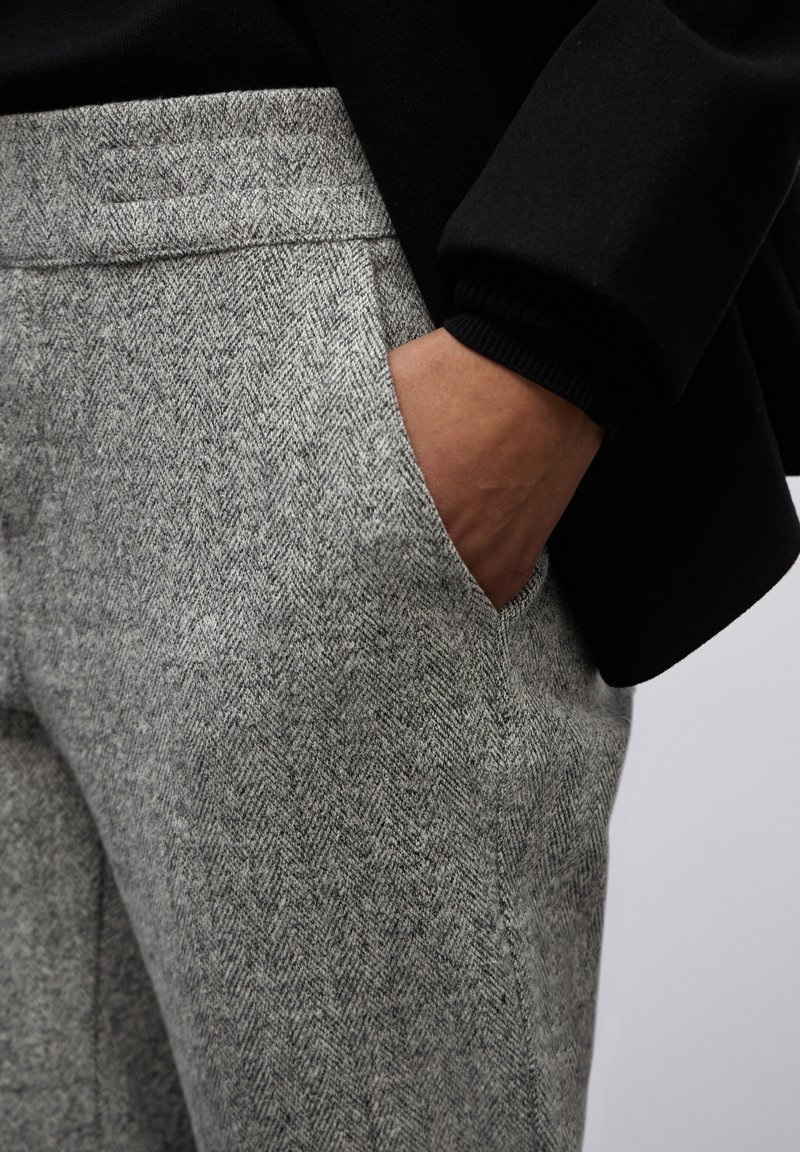 Grijze broek met visgraatpatroon heeft een zijzak en een gladde textuur, gecombineerd met een zwarte kledinghoes op de achtergrond.