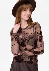 Vrouw met een bruine hoed met een brede rand en een doorschijnende top met lange mouwen met donkere bloemenpatronen, kijkt zijwaarts met een lichte glimlach.