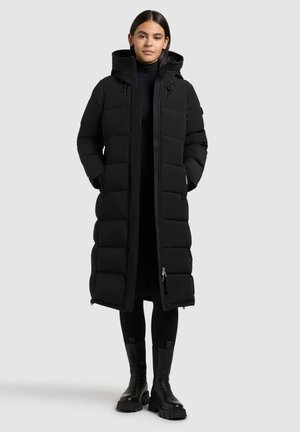 Manteau d'hiver noir long en doudoune avec fermeture éclair et pression, capuche et design matelassé. Porté par-dessus une tenue noire, avec poches et tissu texturé.