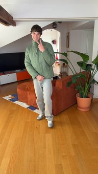 Zelená péřová bunda, světlé džíny a stříbrné tenisky. Hnědá manšestrová pohovka, dřevěná podlaha a velká květina v květináči v pozadí.