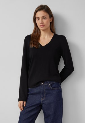 Jonge vrouw met lang bruin haar, gekleed in een zwarte V-hals top met lange mouwen en blauwe spijkerbroek, staand tegen een lichtgrijze muur.