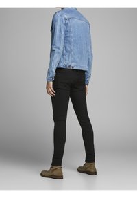 Persona con chaqueta de mezclilla azul claro, pantalones ajustados negros y botines de ante marrón de tobillo de pie contra un fondo gris, de espaldas al espectador.