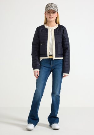 Marineblaue Steppjacke mit cremefarbenem Besatz, kombiniert mit weißem Oberteil und blauen ausgestellten Jeans. Akzentuiert mit einer Leopardenmuster-Mütze und weißen Sneakern.
