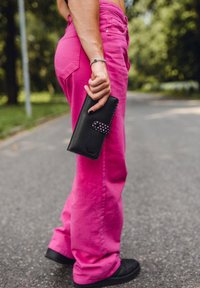 Black leather wallet with a patterned pink clasp held in a hand, paired with bright pink wide-leg pants and black shoes. Background is a road.