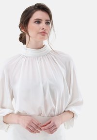 Blusa bianca fluente con un alto colletto arricciato, maniche ampie e una morbida trama di tessuto, abbinata a un pantalone bianco aderente.