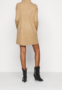 Robe-pull en tricot beige avec col montant, silhouette ajustée et texture côtelée. Associée à des bottines noires à talons.