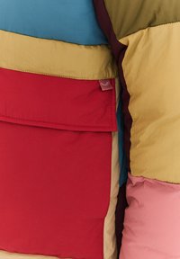 Vícebarevná bunda s částmi v modré, zelené, červené, žluté a růžové barvě. Měkká, nadýchaná textura s viditelnou kapsou a štítkem.