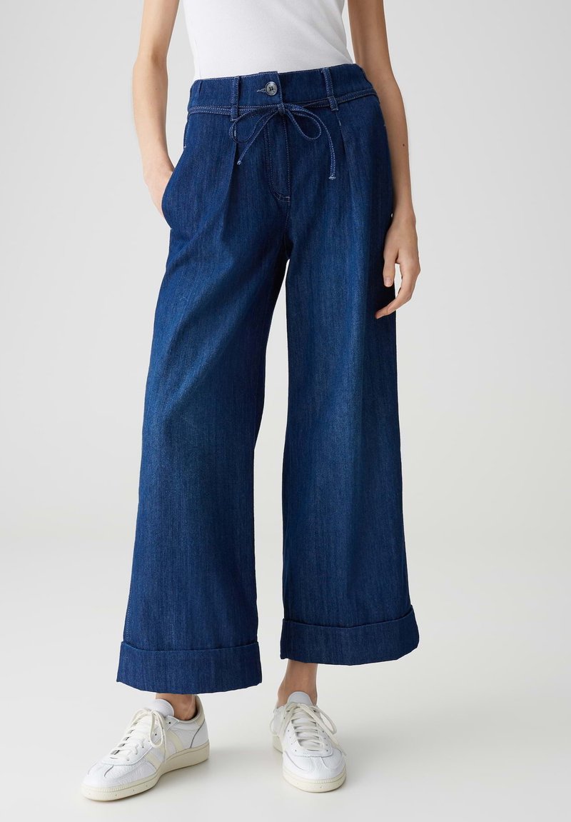 Weite, dunkelblaue Jeanshose mit Knopf- und Kordelzugverschluss, ausgestattet mit umgeschlagenen Säumen und Vordertaschen. Kombiniert mit weißen Sneakern.