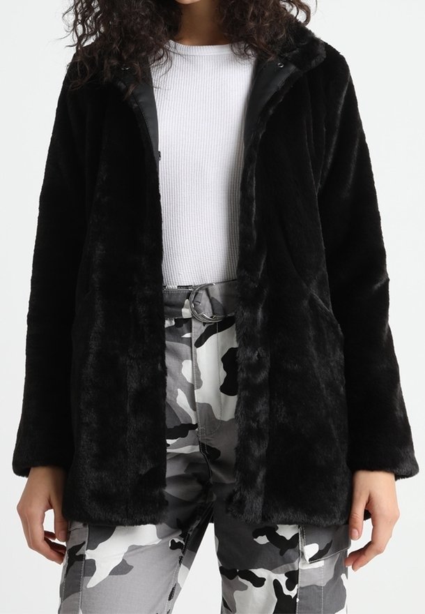 Sort faux fur jakke med hætte, åben front og blød tekstur. Båret over en hvid ribbet bluse og parret med camouflagebukser.
