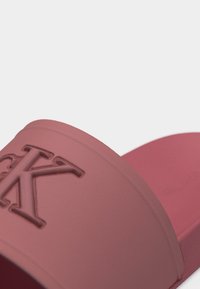 Κοντινό πλάνο ενός ροζ σαγιονάρα με αμμοχρώμα, με ανάγλυφο λογότυπο "CK" στο φαρδύ λουράκι.