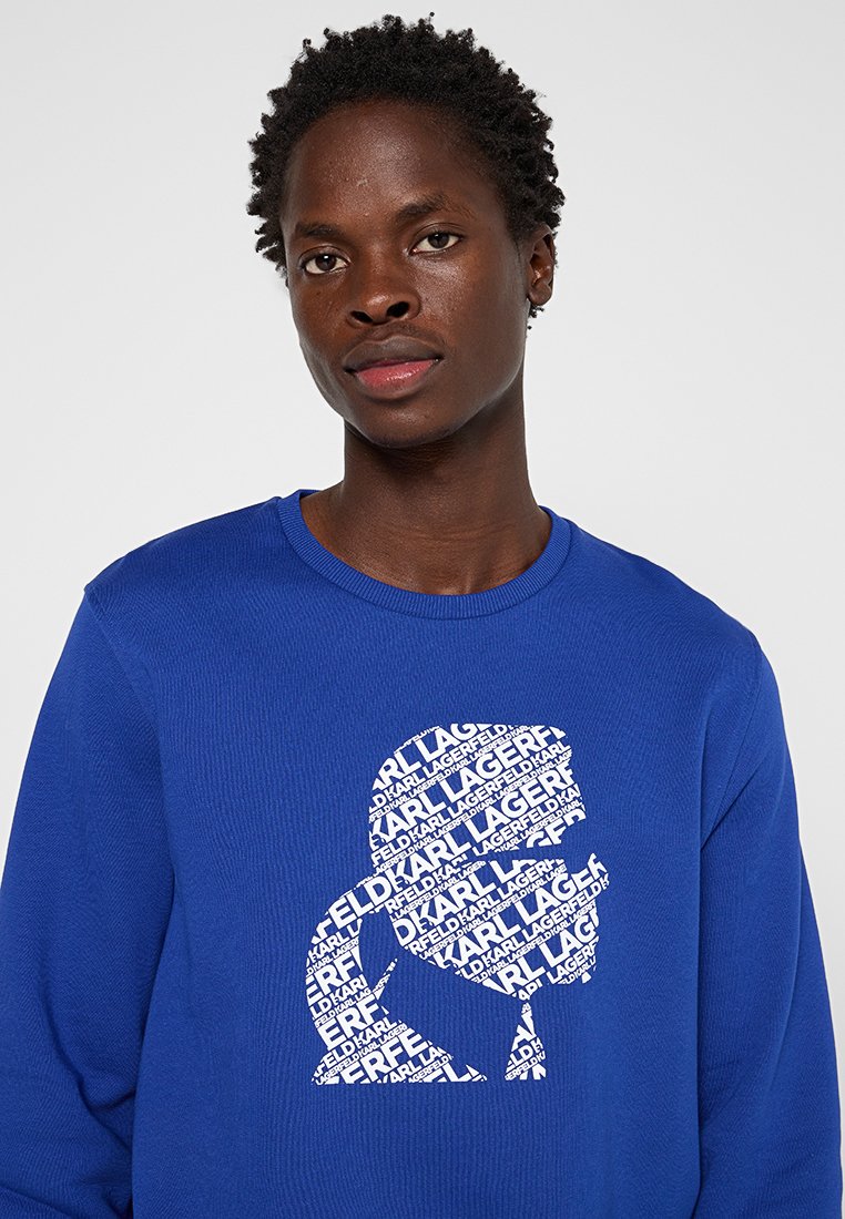 Sweat-shirt bleu avec texture côtelée présentant une silhouette graphique blanche composée de texte répété, mettant en avant un design audacieux et moderne.