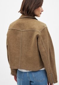 Giacca corta in suede marrone con cuciture in vista, collo a scialle e dettagli con zip sui polsini, abbinata a jeans azzurri chiari.