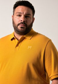 Žluté polo tričko s přední částí na tři knoflíky, jemně texturovaná látka a malé embosované logo na hrudi. Klasický střih s krátkými rukávy.
