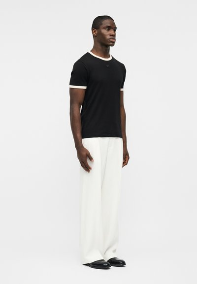 Νεαρός άνδρας όρθιος, φορώντας μαύρο T-shirt με λευκή τρέσα, φαρδιά λευκά παντελόνια και μαύρα παπούτσια, σε απλό φωτεινό φόντο.