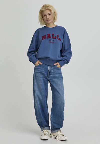 Sweat-shirt bleu avec le texte "BALL 1974" en rouge, porté avec un jean ample bleu clair et des baskets blanches aux accents rouges.
