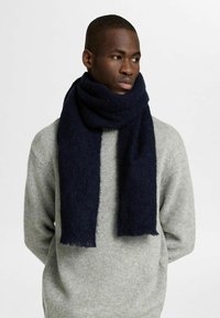 Sciarpa blu navy realizzata in morbido materiale a maglia, con un design spesso e texture, appesa in modo svogliato attorno al collo, con bordi frangiati.