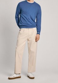 Man som bär en blå tröja, beige högmidjade plisserade byxor och vita sneakers med bruna sulor, stående mot en neutral bakgrund.