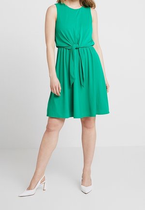 Kobieta w zielonej, bezrękawnikowej sukience o długości do kolan z wiązaniem w talii, w połączeniu z białymi, szpiczastymi sandałami na obcasie ze zapięciem z tyłu, stojąca na jednolitym tle.