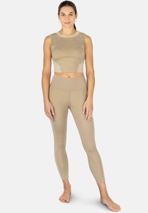 Frau steht barfuß, trägt ein beiges ärmelloses Crop-Top und passende High-Waist-Leggings vor einem einfarbigen Hintergrund.