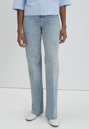 Person, die hellblaue Jeans mit geradem Bein, weiße Schuhe und ein hellblaues Hemd mit Knöpfen trägt und vor einem einfarbigen Hintergrund steht.