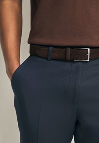 T-shirt en coton marron associé à un pantalon texturé bleu foncé, maintenu par une ceinture en daim marron avec une boucle en métal argenté.