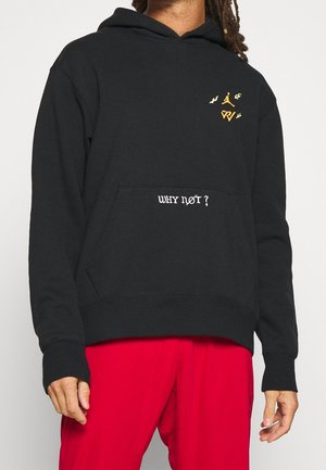 Person trägt einen schwarzen Hoodie mit dem Schriftzug "WHY NOT?" auf der Tasche und kleinen goldenen Logos, kombiniert mit leuchtend roten Hosen.