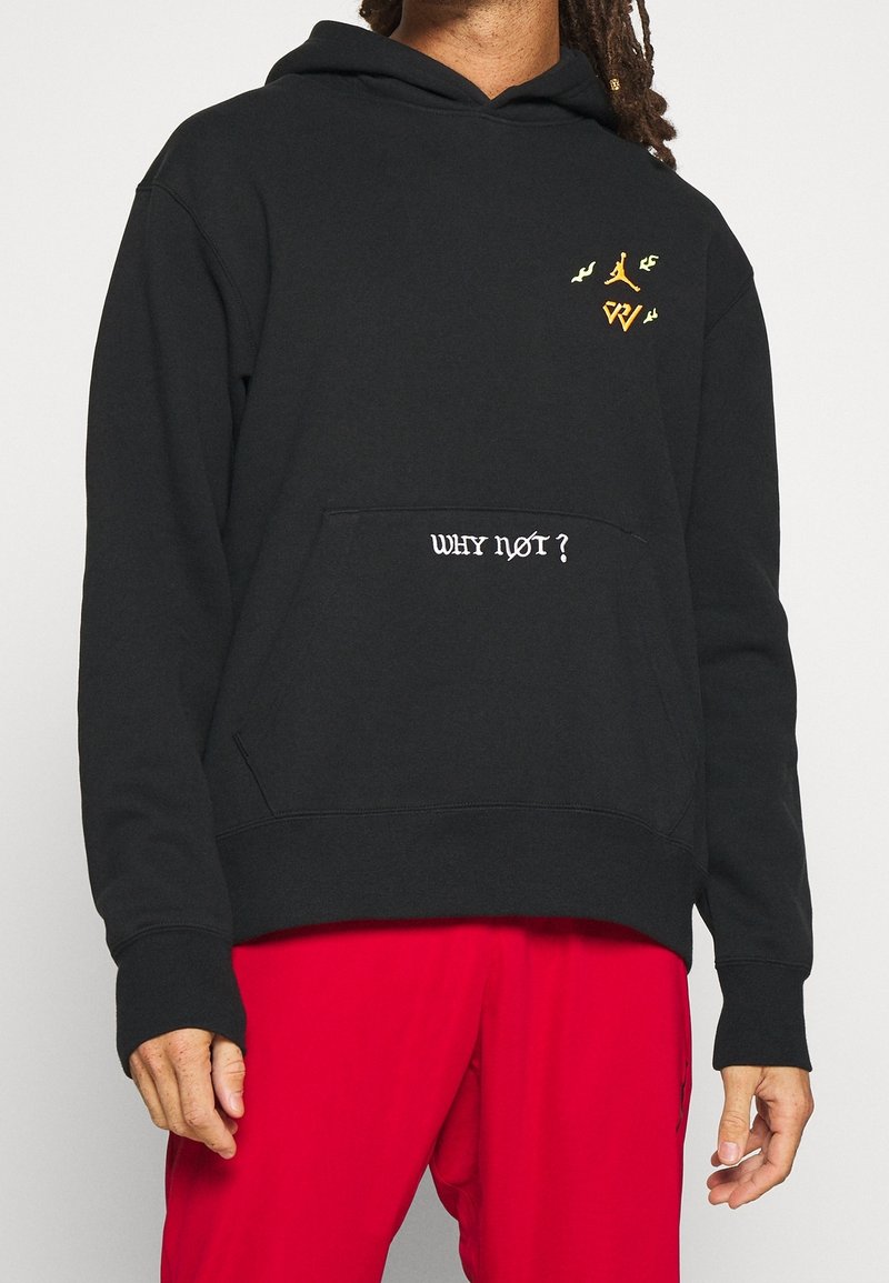 Personne portant un sweat à capuche noir avec le texte « WHY NOT ? » sur la poche et de petits logos dorés, associé à un pantalon rouge vif.