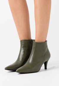 Olivgrüne Leder-Stiefeletten mit spitzer Zehenpartie, seitlichem Reißverschluss und schlankem, niedrigem Absatz, mit glatter Textur und minimalen Details.