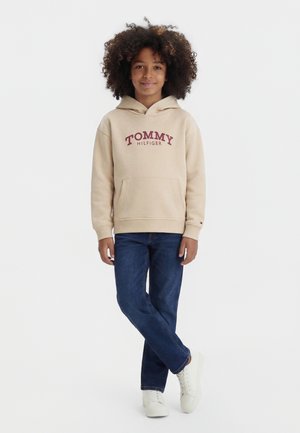 Vaikas garbanotais plaukais, dėvintis smėlio spalvos Tommy Hilfiger džemperį su gobtuvu, mėlynas džinsus ir baltus sportinius batelius, stovintis sudėjus kojas kryžmai.