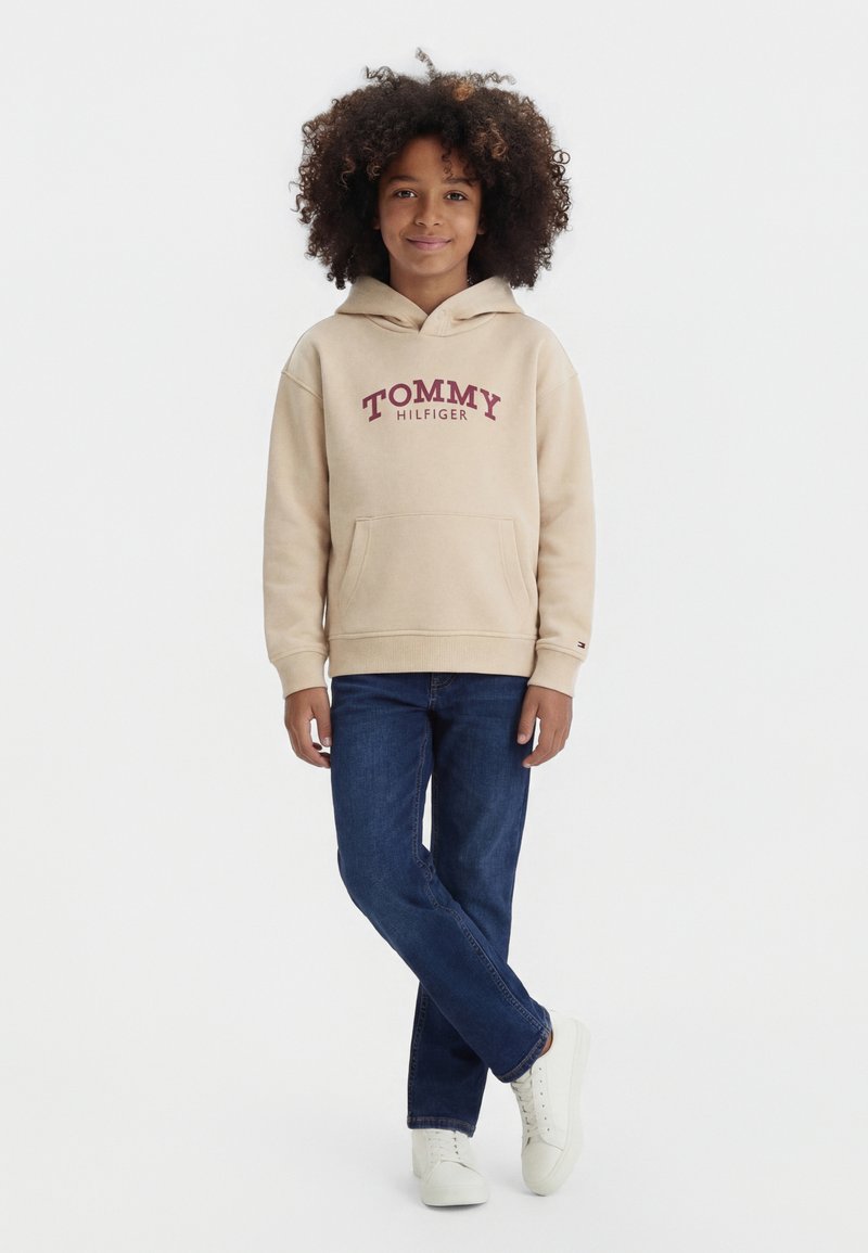 Barn med krøllet hår iført beige Tommy Hilfiger hættetrøje, blå jeans og hvide sneakers, stående med benene krydsede.