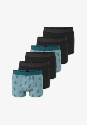 Junger Mann mit lockigem Haar, der graue Boxershorts mit einem türkisfarbenen Ananas-Muster trägt, vor einem schlichten weißen Hintergrund stehend.