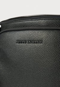Čierna textúrovaná kožená taška so zapínaním na zips a debosovaným logom "Armani Exchange". Vyznačuje sa hladkým a odolným materiálom.