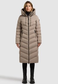 Beige dunjakke til knæene med chevronmønster, hætte, lynlåsfrem og sidelommer; båret over sorte bukser og støvler.