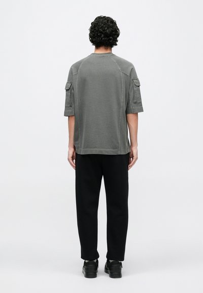 Šedé tričko s krátkými rukávy a kapsami ve stylu cargo, doplněné černými kalhotami a černými botami, pohled zezadu.