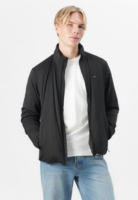 Svart zip-up jakke med høy krage, lett materiale og subtil logo-detalj på brystet, brukt over en hvit T-skjorte.