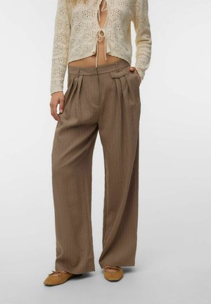 Femme portant un pantalon large marron à fines rayures, un cardigan beige en crochet noué devant, et des ballerines couleur fauve, debout avec une main dans la poche.