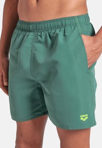 Uomo che indossa dei pantaloni da bagno Arena verdi con cintura elastica e tasca laterale, con la mano appoggiata all'interno della tasca, su uno sfondo bianco.