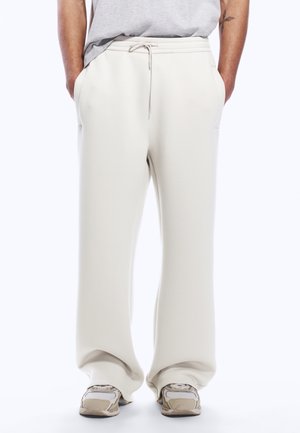 Person som har på seg løse, lyse beige joggebukser med snor og beige sneakers, hender i lommene, mot en ensfarget hvit bakgrunn.