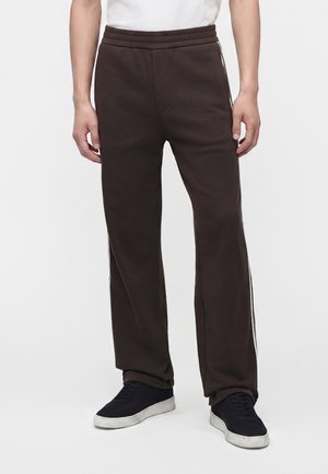Homme portant un pantalon de survêtement marron foncé et des baskets noires aux semelles blanches, debout, les bras détendus le long du corps sur fond blanc.