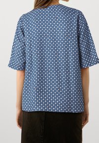 Blusa blu a fantasia con maniche corte. Presenta forme diamantate in bianco e accenti rossi, realizzata in tessuto leggero. Collo rotondo semplice.