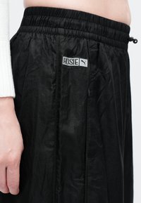 Detailný záber čiernych športových šortiek Puma s nálepkou "ROSIE", ktoré má na sebe osoba s bielym rebrovaným rukávom viditeľným na ľavej strane.