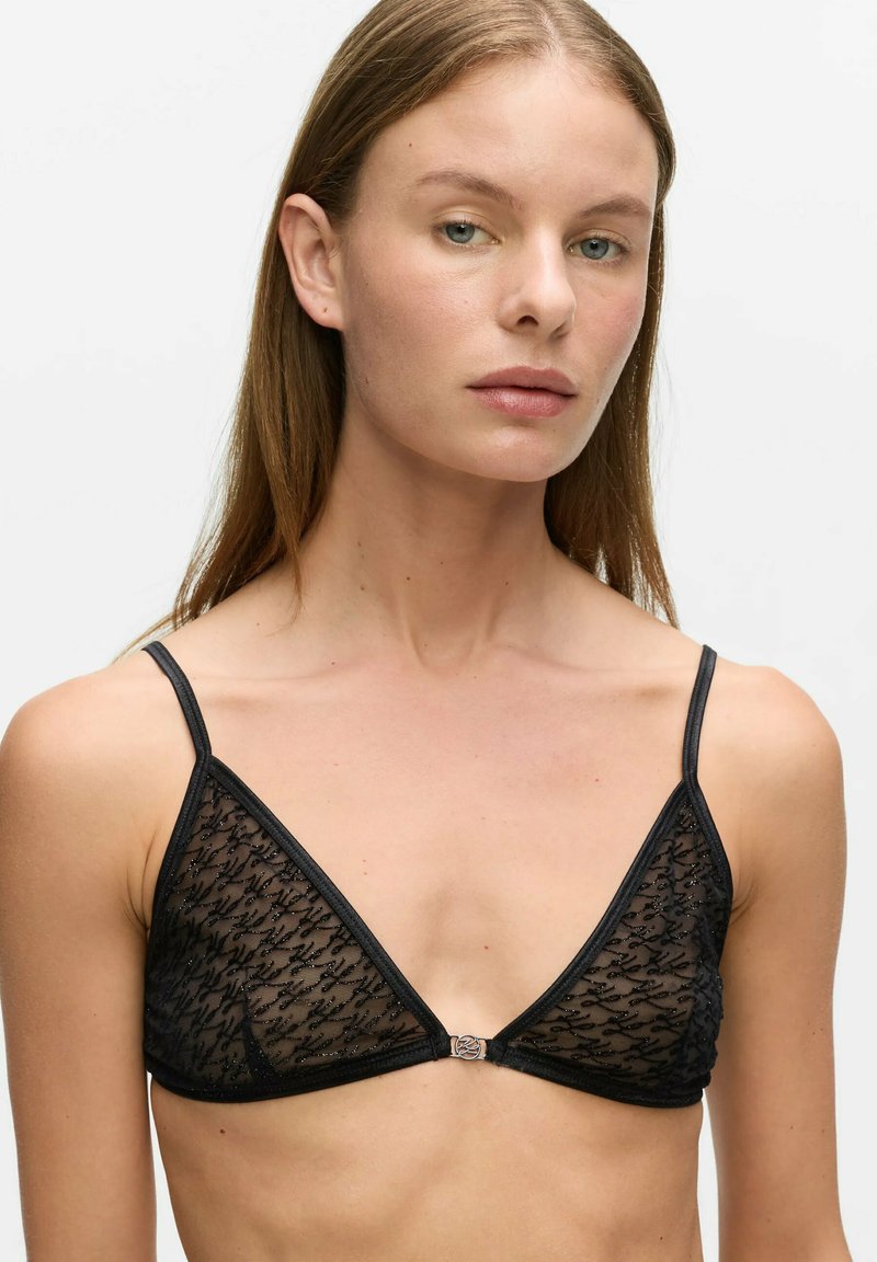 Jeune femme aux cheveux bruns et raides porte un bralette en dentelle noire transparente avec fines bretelles et petite fermeture frontale sur fond uni.