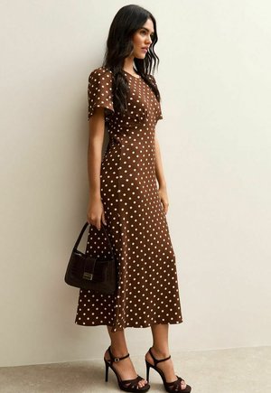 Kobieta w brązowej sukience midi w białe kropki, trzymająca ciemnobrązową torebkę i nosząca czarne sandały na wysokim obcasie.