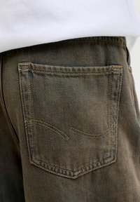 Smeđe traperice prikazuju stražnji džep s detaljima šivanja. Tkanina se odlikuje teksturiranim, rebrastim uzorkom i žutim nitima.